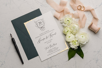 Elegant wedding invitation with gold foil edges, green envelope, white flowers, rings, and ribbon.