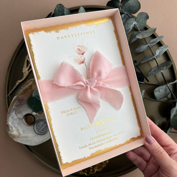 Elegant wedding invitation with gold foil, blush pink ribbon, and floral detail in a gift box.